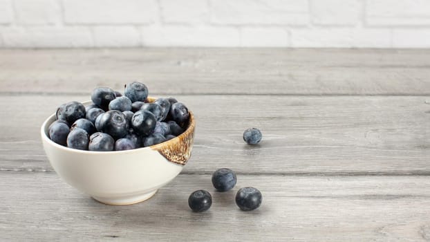 Small bowl full of blueberries, some of them spilled on gray wood desk.