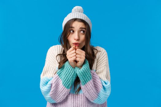 Silly cute, lovely young woman in winter hat, sweater, shaking from cold, blowing hot air at hands to warm-up after playing snowballs outdoors, standing innocent and tender over blue background.