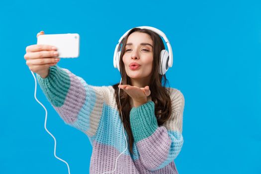 Romance, winter holidays, women concept. Attractive, sensual and flirty young brunette woman in winter sweater, wearing big headphones, holding smartphone and blowing kiss at phone.