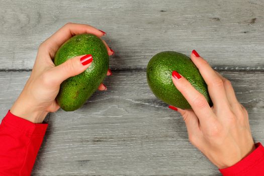 Table top view on woman hands with red nails, holding two green whole avocado over gray wood desk.