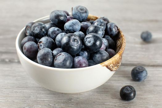 Small bowl full of blueberries, some of them spilled on gray wood desk.