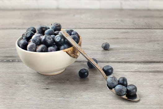 Small bowl full of blueberries, with spoon and some spilled berries on the gray wood desk.