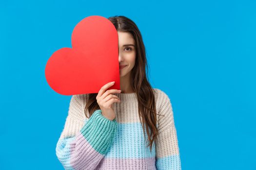 Women, romance and valentines day. Romantic cute, dreamy girl in winter sweater, cover half of face with red big heart sign, looking camera slightly smiling, confessing love, blue background.
