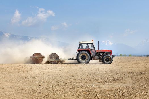 Tractor pulling heavy metal rollers, preparing field in spring, with mountains in background.