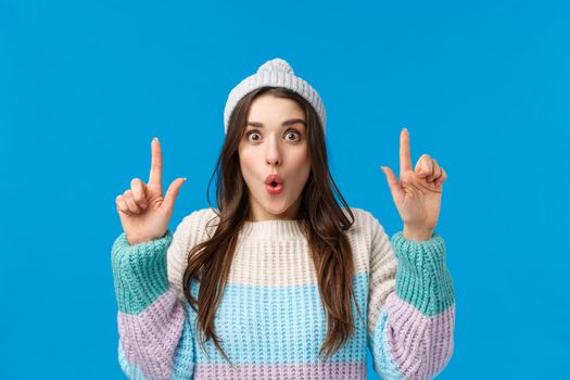 Wow so cool, check this out. Impressed and astonished brunette woman in winter hat, sweater, folding lips amused, looking camera and pointing fingers up, telling about amazing event, blue background.