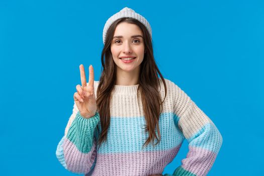 Say cheese. Cute feminine caucasian brunette woman in winter hat and sweater, enjoying christmas holidays, vacation on ski resort, showing peace sign and smiling camera happy, blue background.