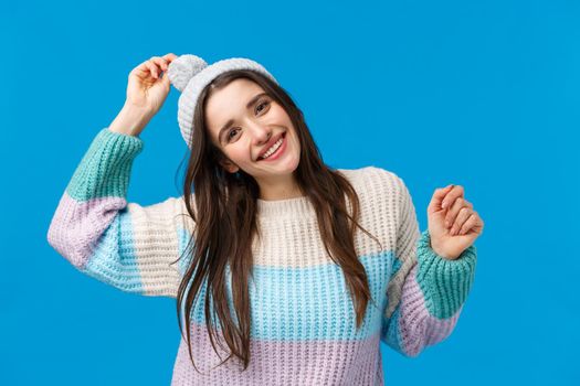 Pretty cheerful and happy, smiling young woman celebrate christmas, winter holidays, dancing and having fun on ski resort, mountains, wearing hat and sweater, standing happy over blue background.