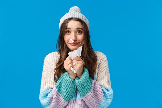 Silly and cute lovely, smiling brunette girl in winter sweater, hat, have runny nose, caught cold on winter holidays, apologizing for sneezing, holding napkin, standing blue background.