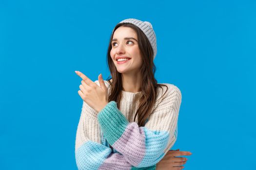 Happy charismatic, friendly young hipster girl in winter hat, sweater, pointing looking upper left corner with pleased smile, feeling well, enjoying new year holidays, standing blue background.