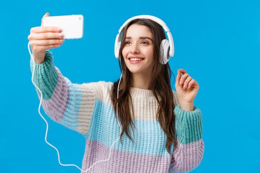 Waist-up portrait cute pretty young woman with long dark hair, cheerful smile, wearing headphones, taking selfie on smartphone, raise hand with phone and posing, standing blue background.