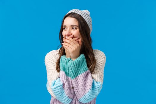 Emotions, fun and happiness concept. Cheerful young caucasian woman in winter sweater, hat, laughing out loud looking aside, laughing close mouth with hands, joking around, standing blue background.