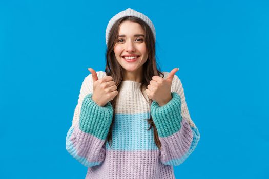 Super good, awesome idea. Cheerful glad cute brunette woman in winter hat, sweater, smiling and showing thumbs-up in approval, encourage friend, say congrats or yes, nod agreement, blue background.
