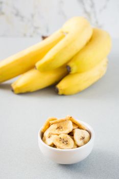 Baked banana chips in a white bowl and a bunch of bananas on the table. Fast food. Close-up. Vertical view