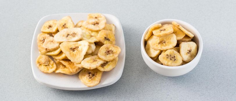 Baked banana chips in a white bowl and saucer on the table. Fast food. Web banner. Close-up