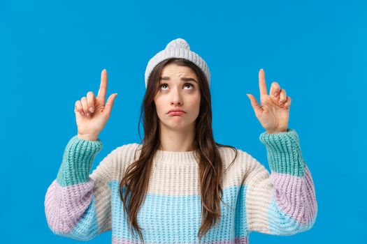 Upset and gloomy, sulking cute caucasian woman in winter hat, sweater, looking pointing fingers up with regret or jealous, envy as cant buy something, desire have it, standing blue background.