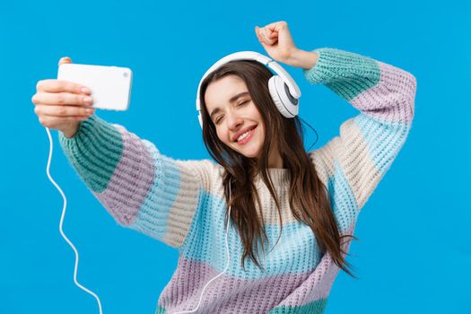 Girl recording video for vlog. Cheerful and carefree happy dancing woman wearing headphones, close eyes and smiling, taking selfie on smartphone during awesome mood, having fun, blue background.