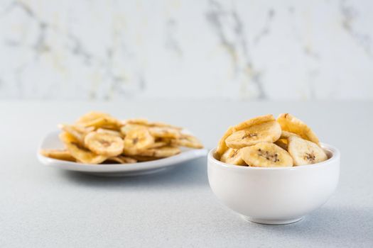 Baked banana chips in a white bowl and saucer on the table. Fast food. Copy space