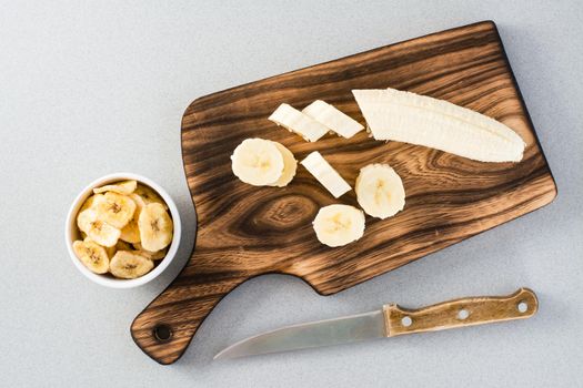 Banana slices on a cutting board and a knife and banana chips in a bowl on the table. Fast food. Top view
