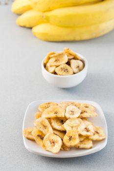 Baked banana chips in a white bowl and saucer and a bunch of bananas on the table. Fast food. Vertical view