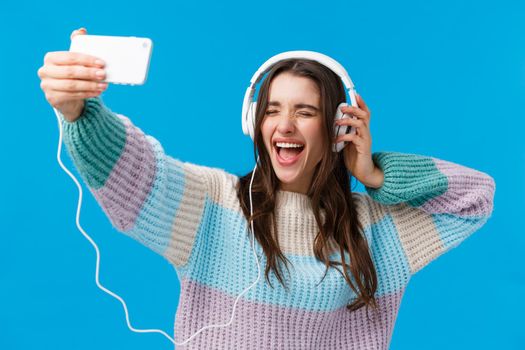 Upbeat, charismatic and happy smiling young gorgeous woman in winter sweater, enjoying awesome music, taking selfie as listening favorite song in headphones, holding smartphone, blue background.