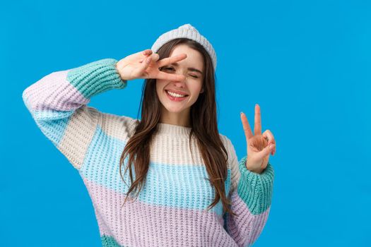 Pack your things and travel. Joyful and carefree upbeat smiling woman, wear winter sweater and hat, wink camera and make peace signs, tilt head, enjoying beautiful, amazing vacation, blue background.