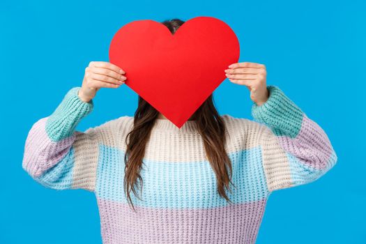 Be my valentine. Cute romantic shy girl in sweater hiding head behind red big heart sign, express love, confessing to friend, want start relationship showing sympathy and affection, blue background.