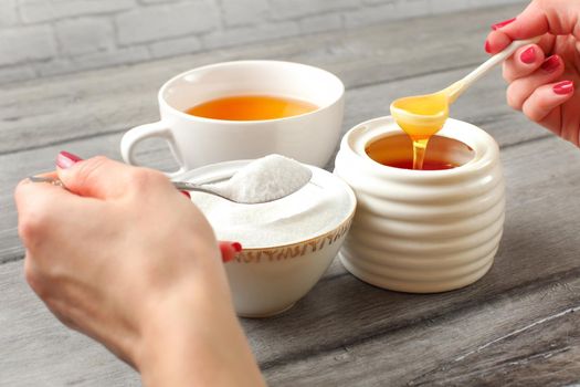 Young woman hands holding spoon of sugar, and honey deciding what to put in tea. Refined vs natural sweetener concept.