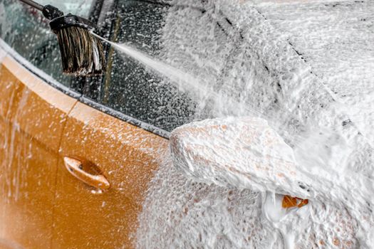 Yellow car side mirror completely covered with thick shampoo foam, more spraying from the brush, when cleaned in carwash.