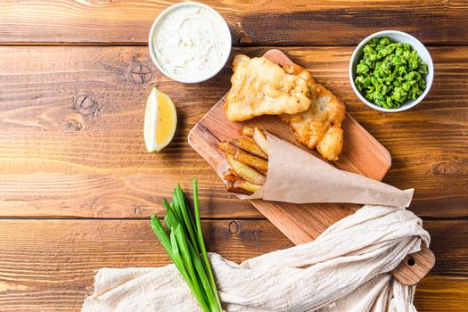 Traditional Fish and chips with mashed peas, tartar sauce inn crumpled paper cone on wood chopping board dip and lemon - fried cod, french fries, lemon slices, tartar sauce, ketchup tomatoe served in the Pub or Restaurant over light old wooden planks table top view space for text.