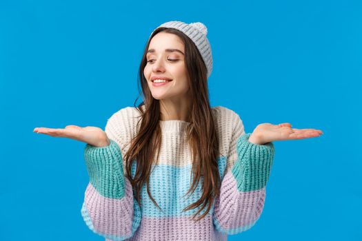 Sensual, cute and tender feminine young woman in sweater, winter hat, raising hands, holding products on palms, smiling making choice, have two good gifts, special discount offers, blue background.