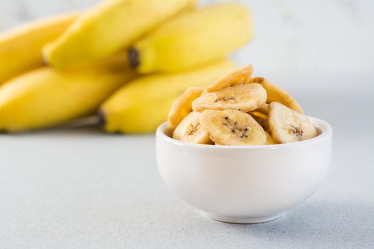 Baked banana chips in a white bowl and a bunch of bananas on the table. Fast food. Close-up