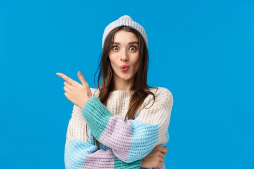 Wow its interesting, lets see. Intrigued and excited cute smiling woman in winter sweater, hat, folding lips curiously pointing finger left, telling about awesome event, standing blue background.