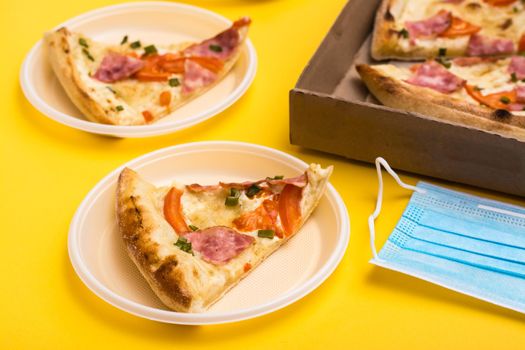 Takeaway and delivery. A slice of pizza in a disposable plastic plate, a box of pizza and a protective mask on a yellow background