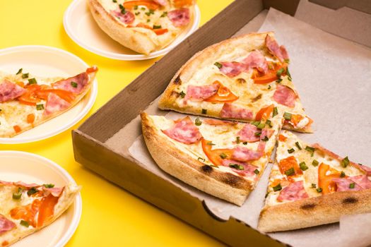 Takeaway and delivery. Pizza in a cardboard box and pieces of pizza laid out on disposable plates on a yellow background