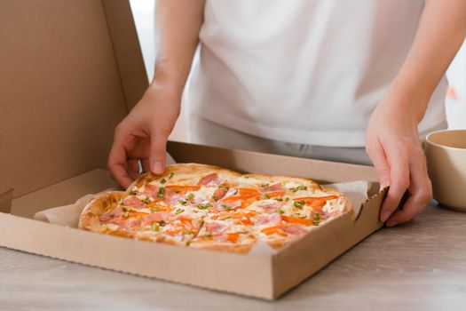 Takeway food. A woman holds a cardboard box with ready-to-eat pizza on the table in the kitchen.