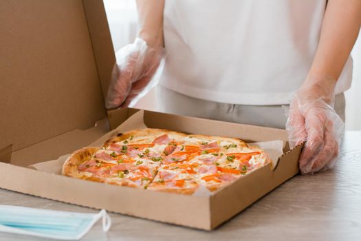Takeaway food. A woman in disposable gloves holds a cardboard box of pizza and a protective mask on the table in the kitchen
