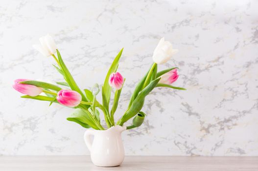 A bouquet of white and pink tulips with green leaves stands in a jug on the table. Selective focus. Delicate postcard
