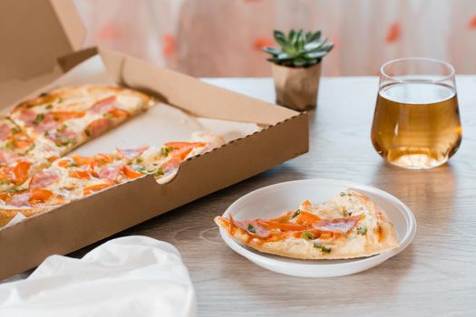 Takeout food. A slice of pizza in a disposable plastic plate, beer and a box of pizza on the table in the kitchen.