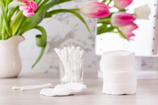 Personal hygiene, cleanliness and skin care. Cotton pads and swabs in a glass on a table in front of a mirror and a bouquet of tulips in a vase