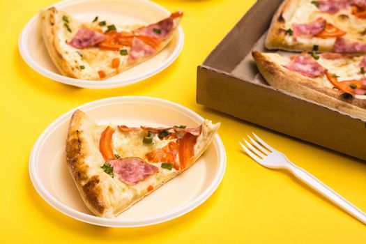 Takeaway and delivery. Pizza slice in disposable plastic plate, pizza box and fork on yellow background