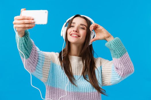 Attractive, sassy and stylish flirty brunette woman in winter sweater, taking selfie using smartphone, showing heart sign and smiling phone camera, wearing headphones, blue background.