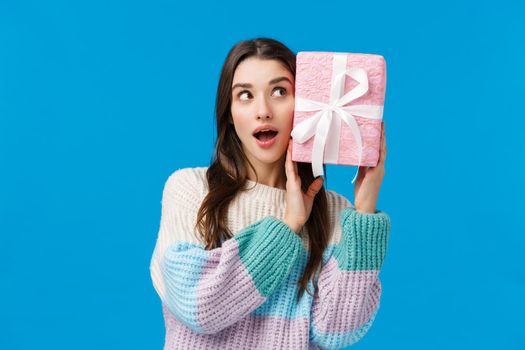 Waist-up portrait curious attractive young woman 20s, brunette shaking cute present to guess what inside, feeling intrigued receiving christmas gift, standing wondered and amused blue background.