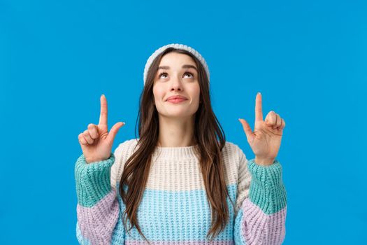 Intrigued and cute caucasian woman with long dark hair, wear winter sweater, hat, looking pointing up with satisfied curious smile, found interesting product, watching snowlakes falling sky.