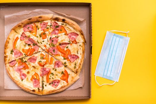 Takeaway and delivery. Ready-to-eat pizza in cardboard box and protective mask on yellow background