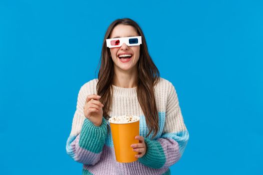 Girl watching comedy in cinema. Carefree happy young brunette woman in winter sweater, enjoying funny movie, laughing out loud eating popcorn, wearing 3d glasses, standing blue background.