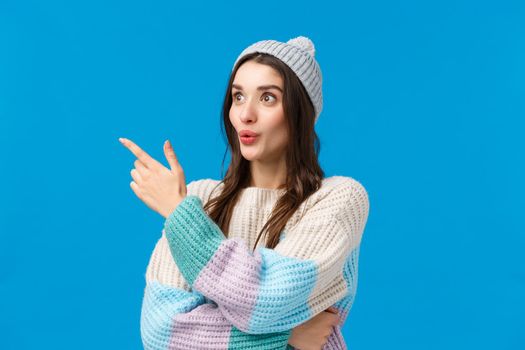 Girls attention caught something interesting happening nearby. Intrigued, curious good-looking brunette woman in winter sweater, hat, looking pointing left with folded lips, amazed, blue background.