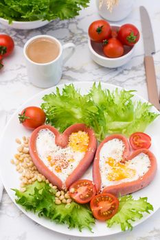 Love breakfast. Fried eggs in heart shaped sausages, lettuce and cherry tomatoes on a plate and a cup of coffee. Vertical view