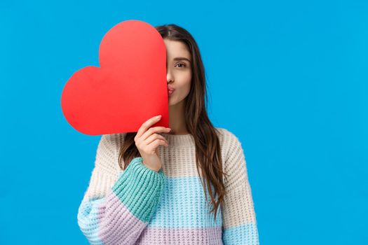 Romantic gesture, love and valentines day concept. Dreamy attractive girlfriend in winter sweater, cover half face with red heart, folding lips in kiss, standing blue background showing sympathy.