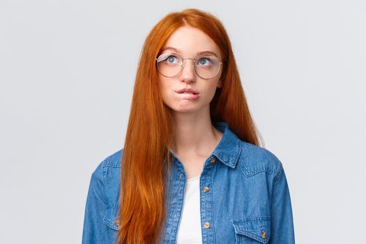 Thoughtful and curious cute redhead teenage girl, thinking, biting lip intrigued and looking up as planning something, recall, pondering between choices, standing white background.