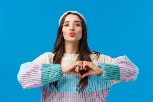 Showing her love. Pretty sensual and cute, coquttish brunette woman in winter sweater, hat, blowing air kiss, folding lips silly and showing heart gesture over chest, show affection and passion.